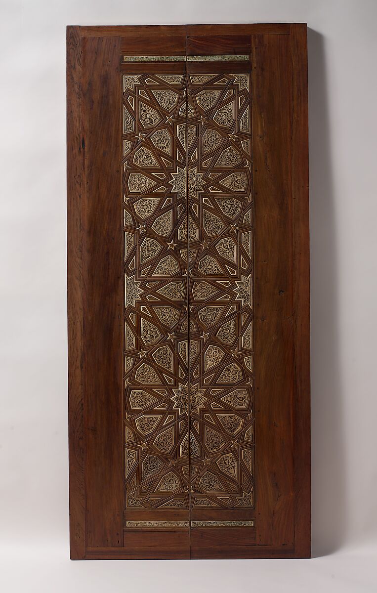 Two tall wooden minbar doors with pale geometric inlay forming interlocking stars and polygons across the paired central panels.